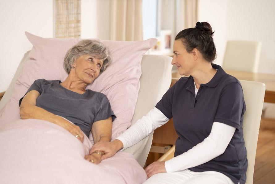 Eine Pflegerin hält die Hand von einer älteren Dame die im Bett lieg