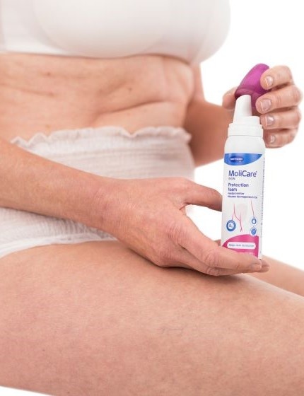An older women siting in her underwear holding up a molicare skin product bottle before applying it.