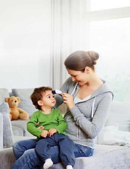 A mother is gently taking her child’s temperature using a veroval thermometer while they sit together on a couch. The scene conveys a sense of care and comfort.